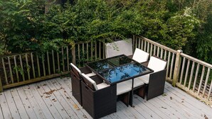 An outdoor deck with a glass-top table and wicker chairs, surrounded by a wooden fence and lush greenery. - The Hawksbill Haus (w/ hot tub) (Waynesboro)