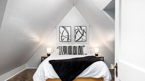 A bedroom with a sloped ceiling, a bed with white bedding, two black and white abstract paintings above the bed, and two bedside lamps. - The Hawksbill Haus (w/ hot tub) (Waynesboro)