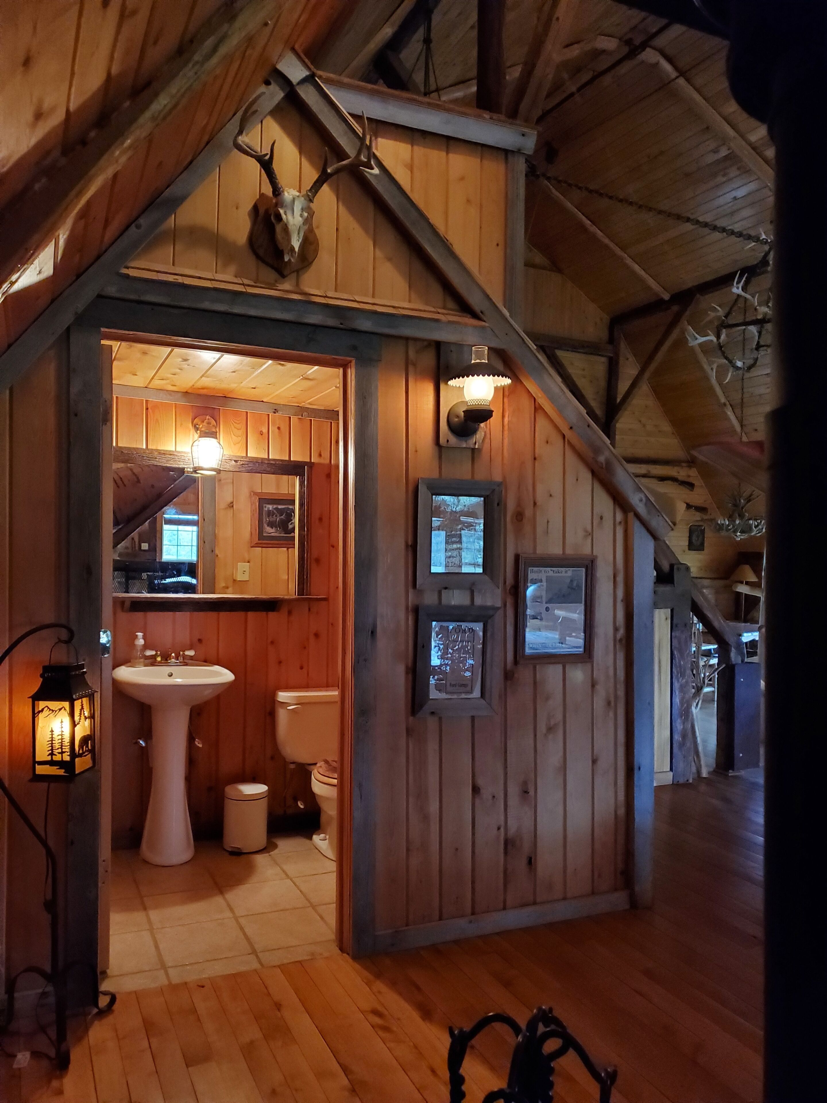 A rustic bathroom with wooden walls, a sink, toilet, and a mirror. There are wall-mounted lights and a lantern.