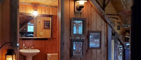 A rustic bathroom with wooden walls, a sink, toilet, and a mirror. There are wall-mounted lights and a lantern.