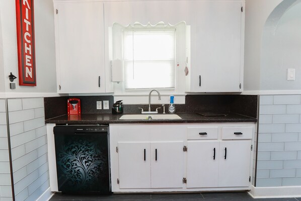 A kitchen with white cabinets, a black countertop, a dishwasher, and a window with blinds. - Cheerful Colonial in Historic Olde Wythe, Hampton, VA (Hampton)