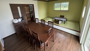 A dining area with a wooden table and chairs, a small kitchen area with a coffee maker, and a window with a view of greenery.