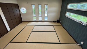 A tatami mat room with a view of a garden through a sliding door, a dark wooden panel wall, and a window with a clock.