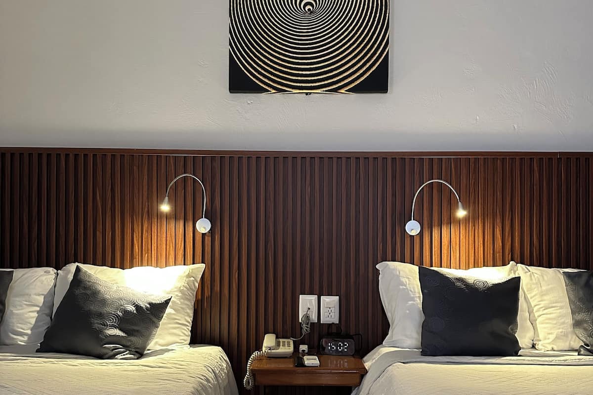 A hotel room with two beds, a wooden panel wall, a wall-mounted artwork, bedside lamps, a nightstand with a phone and clock, and a tiled floor.