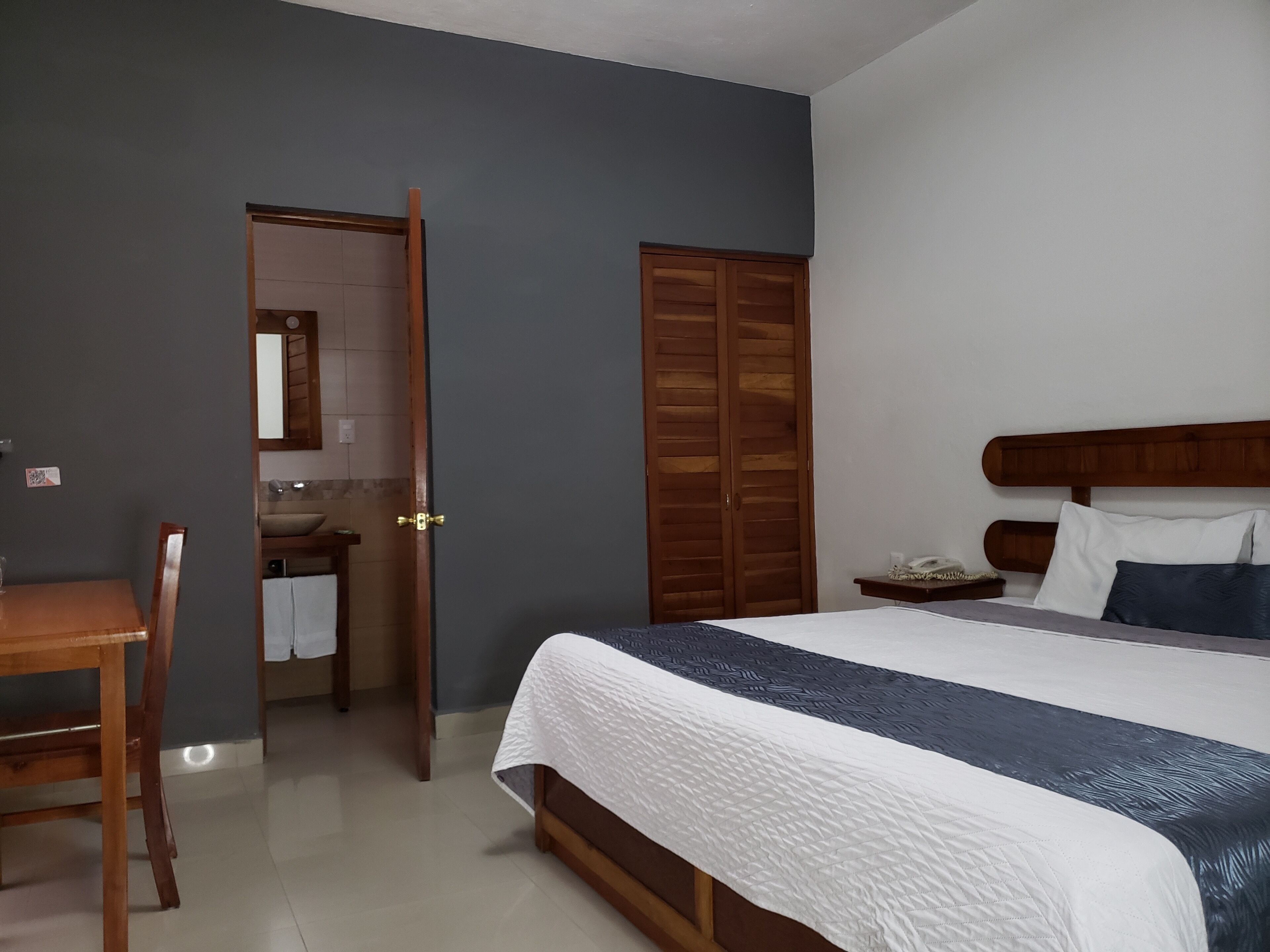 Une chambre à coucher avec un lit, une tête de lit en bois, une table de chevet, un bureau et une salle de bain visible à travers une porte ouverte.