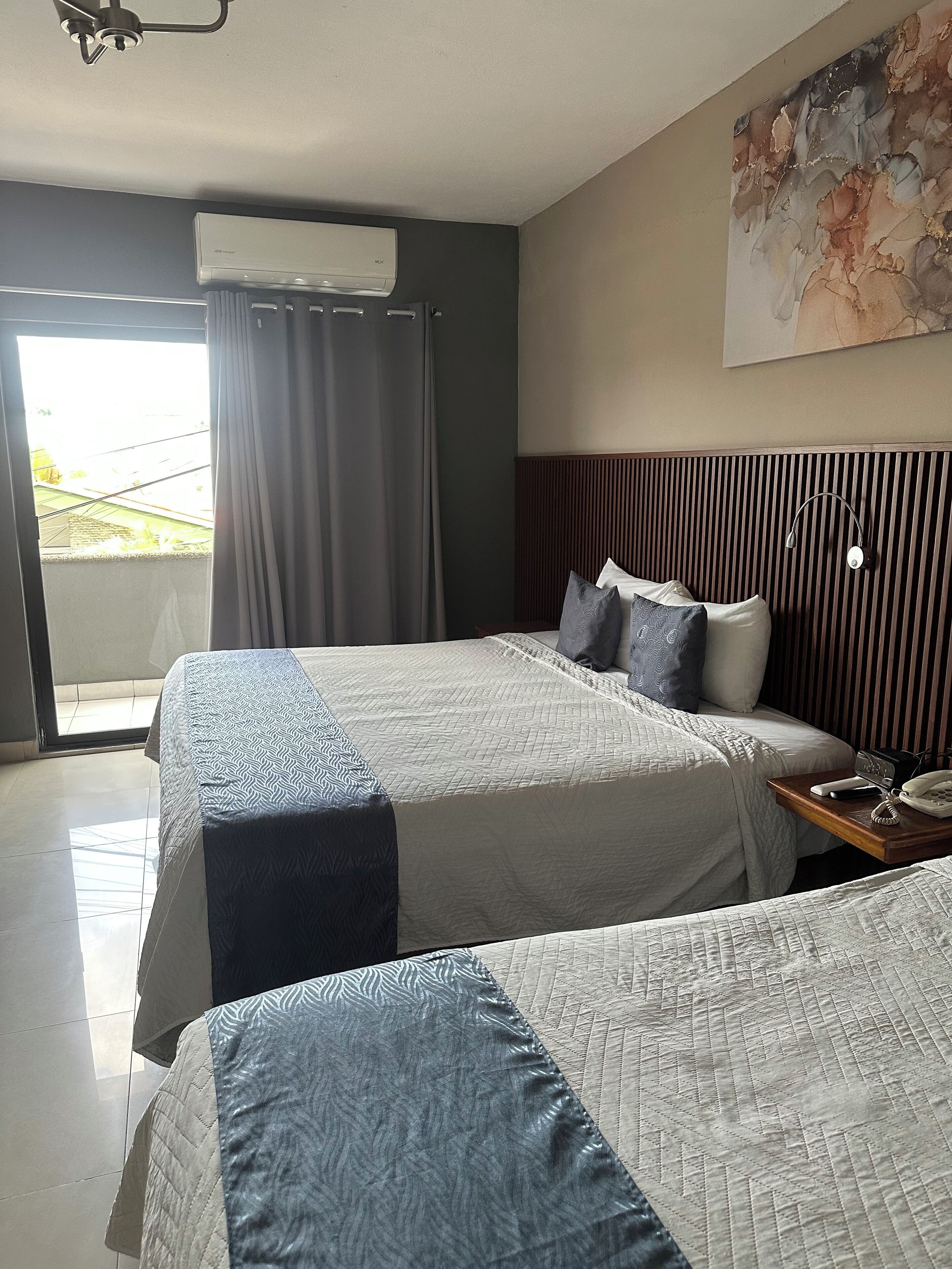 Habitación de hotel con dos camas, una mesita de noche con teléfono y lámpara, un ventilador de techo y un ventanal con cortinas.