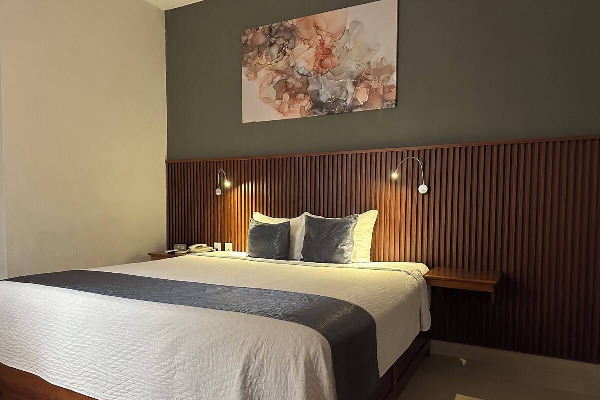 A neatly arranged hotel room with a large bed, a bedside table, a wall-mounted artwork, and a wooden panel headboard.