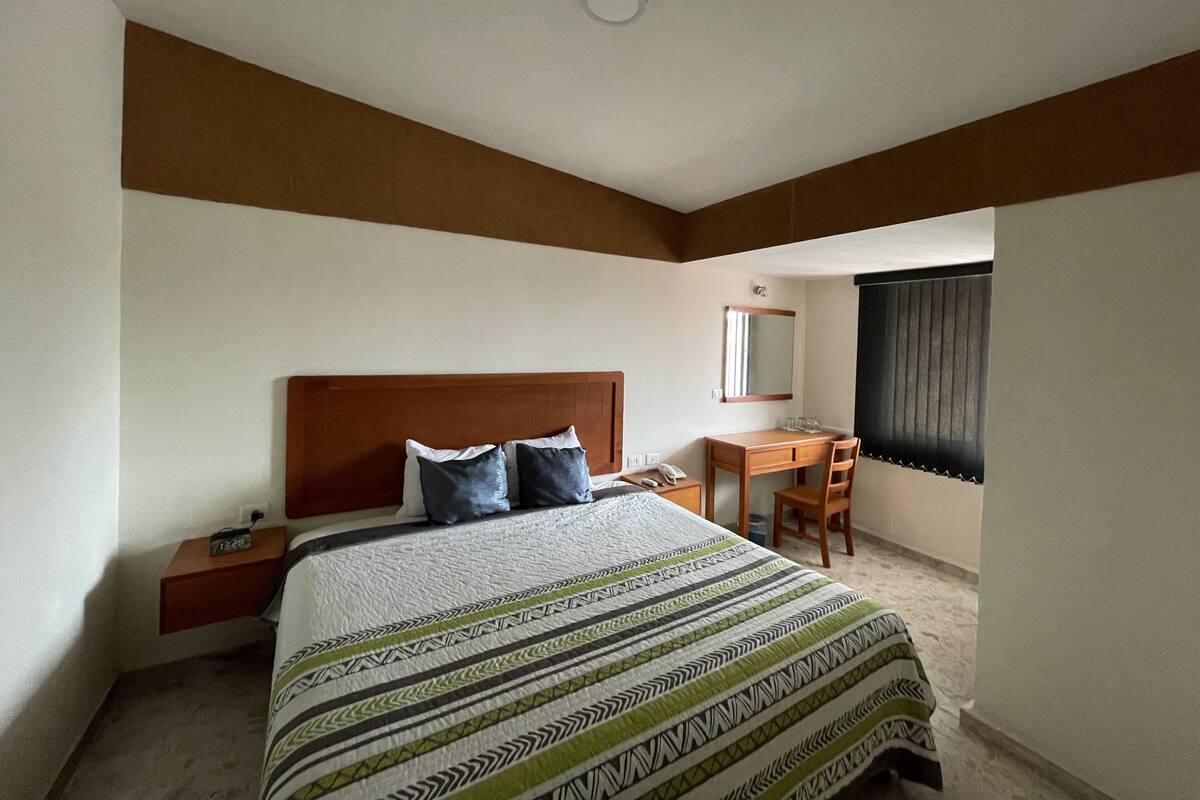 A bedroom with a bed, bedside table, desk, chair, and a window with blinds.