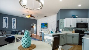 Cocina moderna con armarios blancos, microondas y lavavajillas. Hay una mesa de comedor con una piña como centro de mesa.