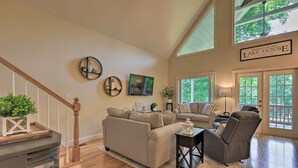 A spacious living room with a high ceiling, a staircase, and a large window offering a view of the outdoors. - Waterfront Eatonton Escape w/ Private Hot Tub! (Eatonton)