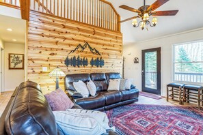 Un salon chaleureux avec un canapé en cuir, un mur en bois orné d’un motif de montagne, un ventilateur de plafond et une grande fenêtre.