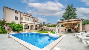 A swimming pool with a swan float, a covered outdoor seating area, and a large house with a tiled roof.