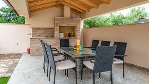 An outdoor patio with a glass-top table surrounded by wicker chairs, a built-in grill, and a stone-tiled floor.