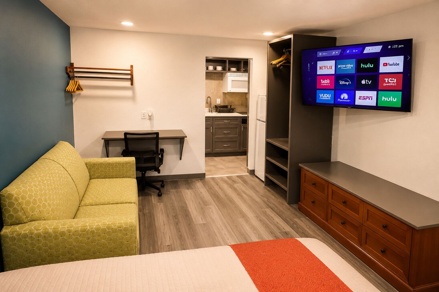 A hotel room with a bed, a desk, a chair, a TV, a wardrobe, and a kitchenette.
