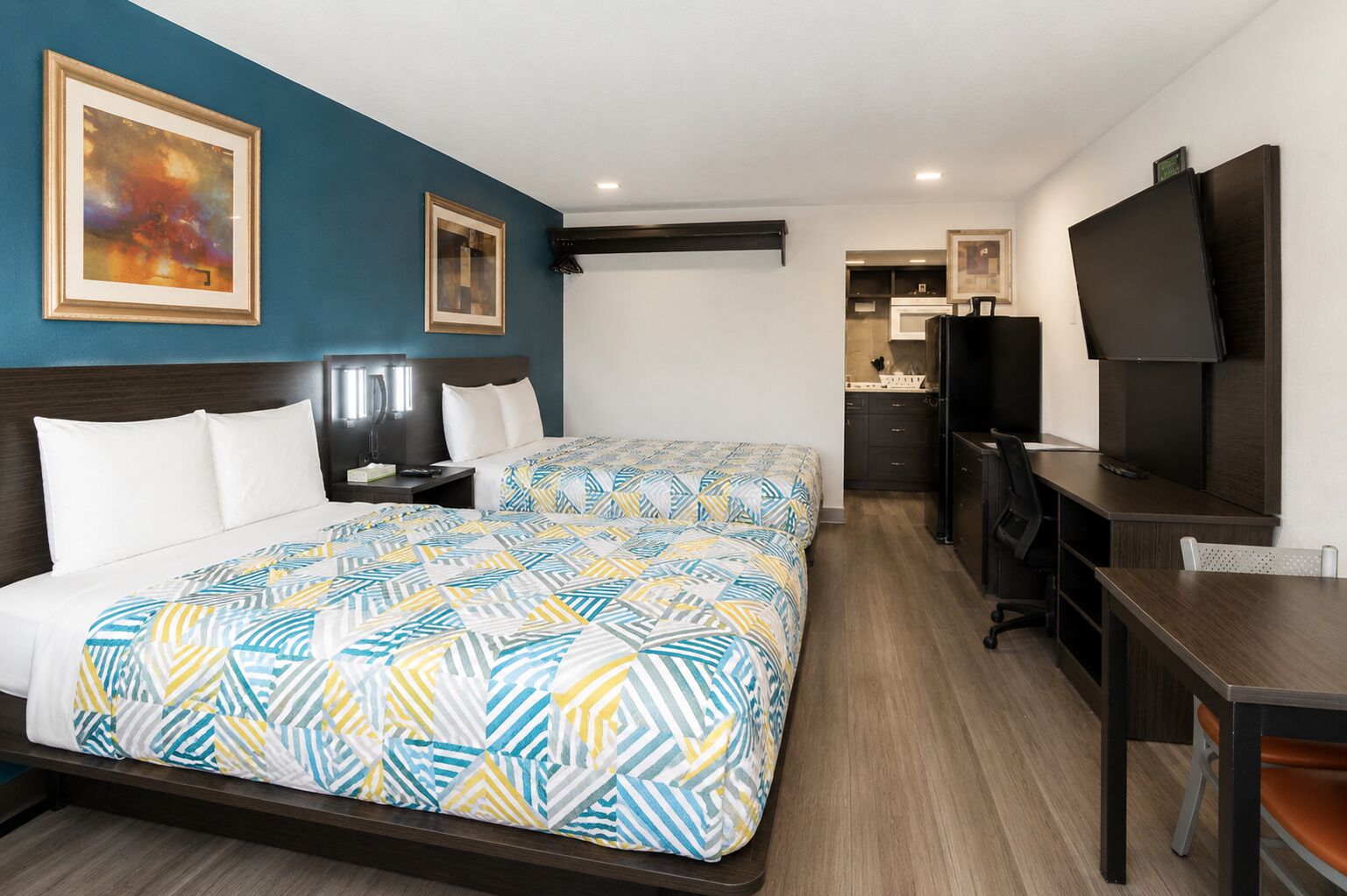 A hotel room with two beds, a desk, a TV, and a kitchenette.