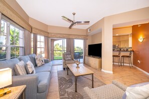 Ein geräumiges Wohnzimmer mit einer Couch, einem Couchtisch und einem Fernseher. Ein Deckenventilator, große Fenster und ein Blick ins Freie sind vorhanden.