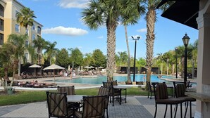 Un espace aménagé au bord de la piscine, avec des chaises longues et des tables, des palmiers et un bâtiment en arrière-plan.