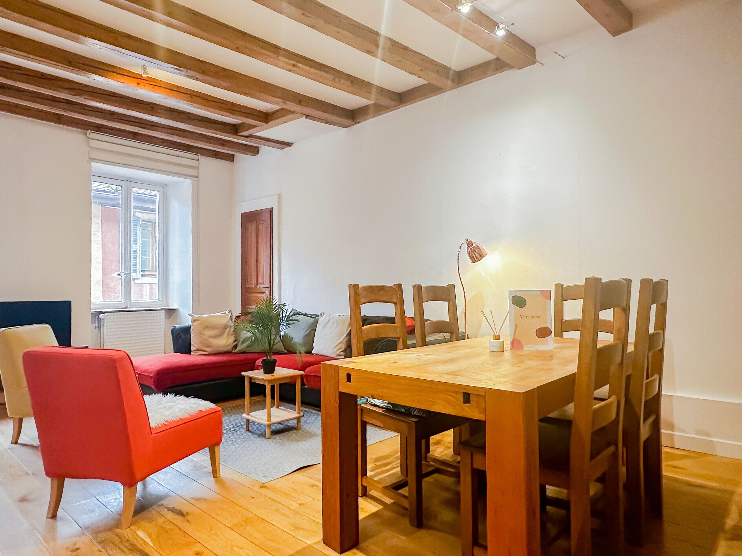 Un coin repas avec une table et des chaises en bois, une petite table d’appoint avec une lampe et une chaise rouge.