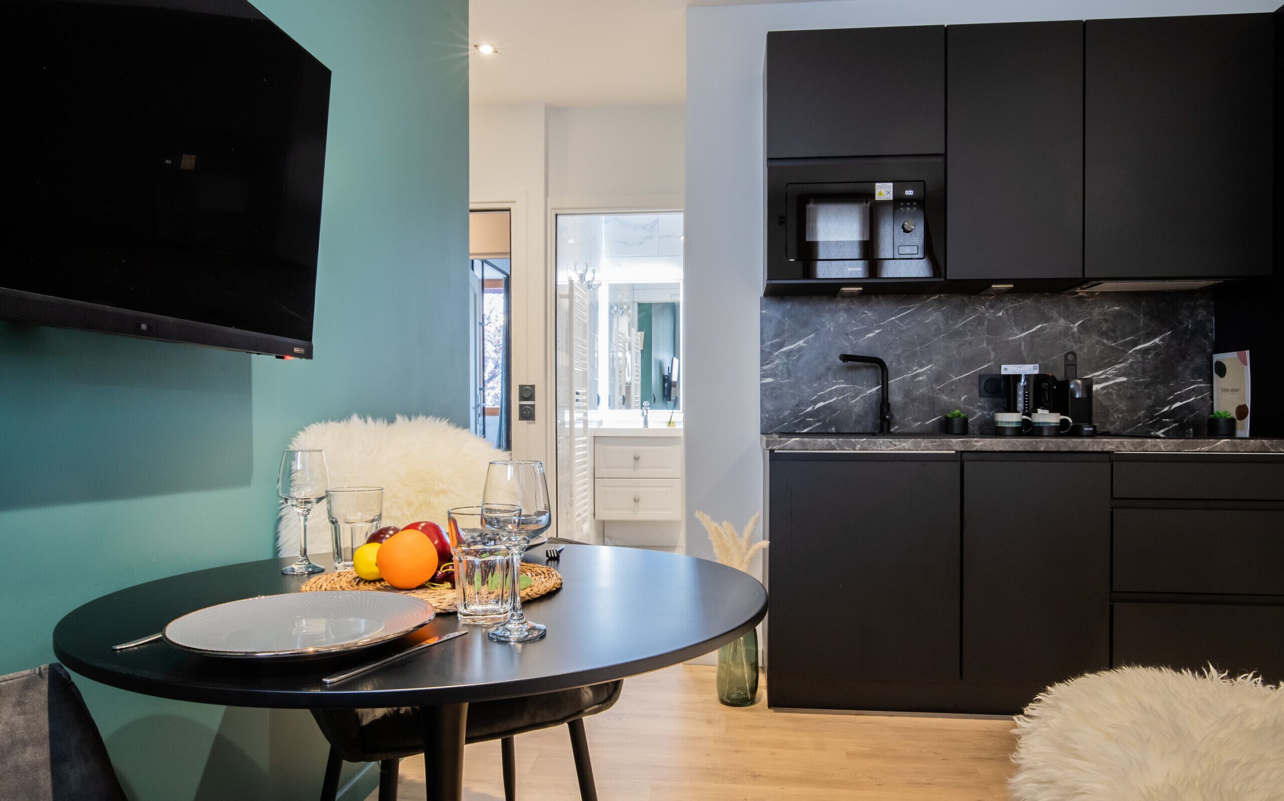 Une cuisine moderne avec des meubles de couleur sombre, un plan de travail en marbre et un coin repas agrémenté d’une table ronde préparée pour deux personnes.
