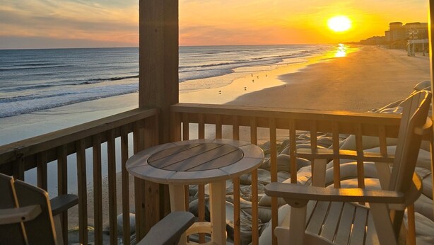 Um deck de madeira com mesa e cadeiras oferece vista para uma praia ao pôr do sol.