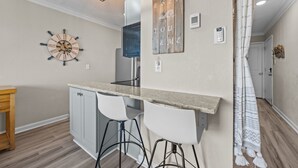 Uma cozinha com ilha central, banquetas, um relógio e um letreiro fixado na parede.