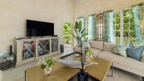 Una sala de estar con televisor en un mueble bajo, un sofá con cojines, una mesa de centro con objetos decorativos y ventanas amplias con cortinas.