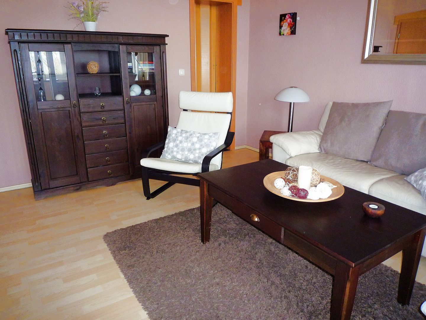 Uma sala de estar com um armário de madeira escura, uma poltrona branca, um sofá e uma mesa de centro com uma bandeja de petiscos.