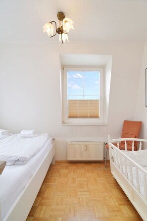 Ein Schlafzimmer mit Holzboden, einem weißen Heizkörper, einem Fenster mit Jalousien und einem weißen Bett mit weißem Kopfkissen.
