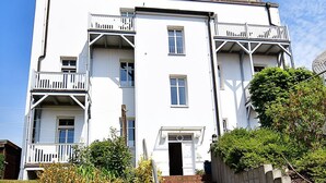Un immeuble blanc à plusieurs étages, doté d’une entrée centrale, de balcons et d’un escalier en briques menant à l’entrée.