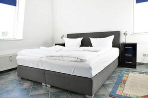 A neatly made bed with white linens, a grey upholstered headboard, and a small bedside table with a lamp.