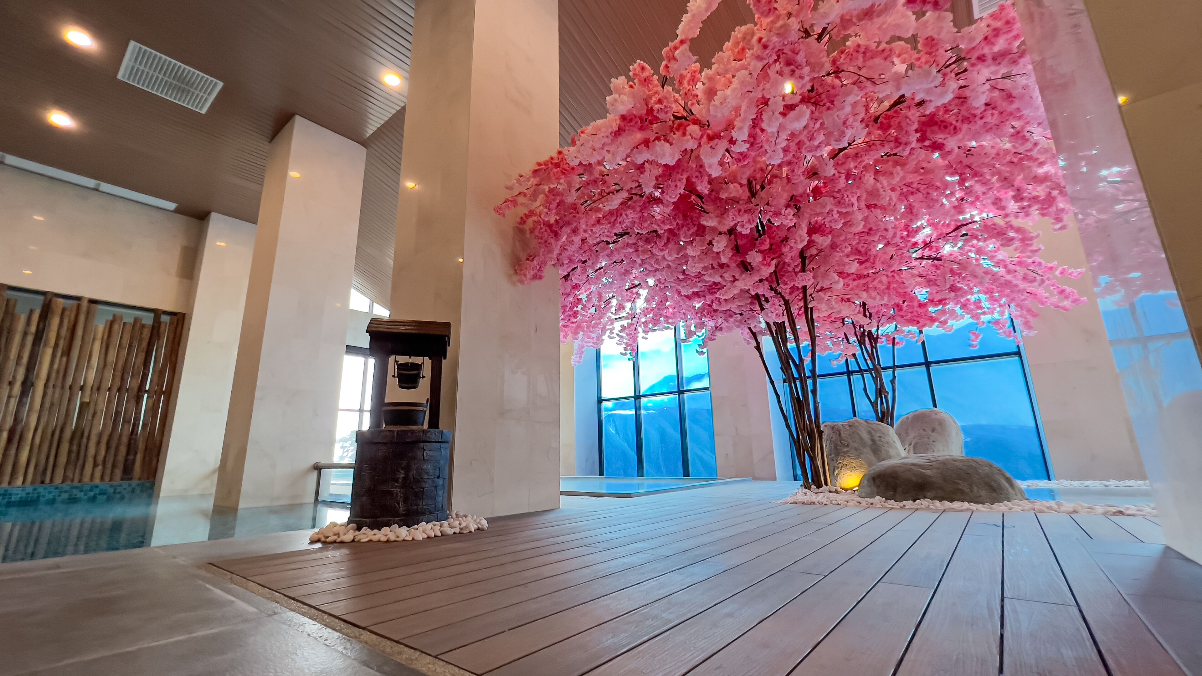 A spacious indoor area with a large artificial cherry blossom tree, a stone water feature, and a view of the outdoors through large windows.