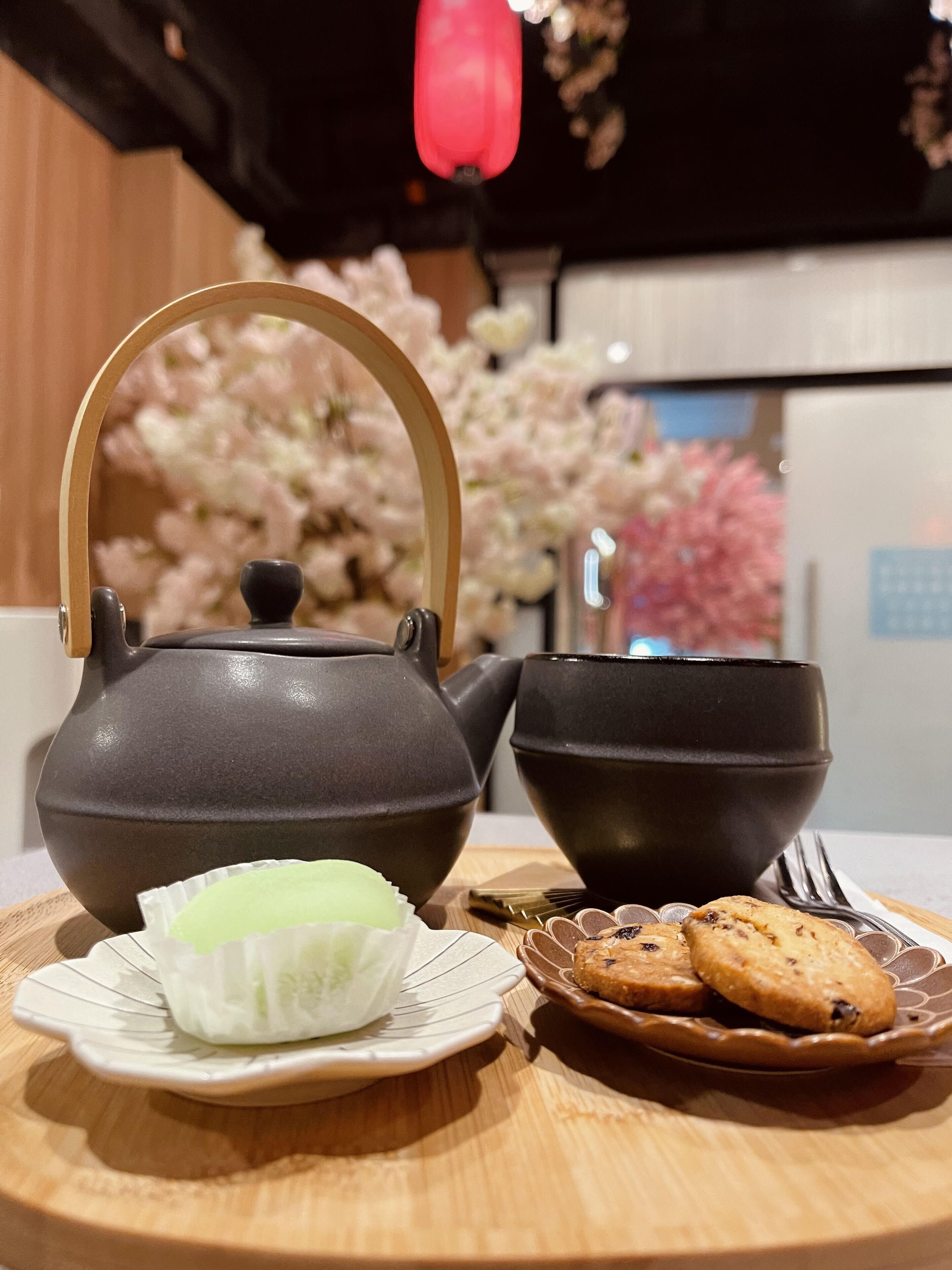A tea set with a teapot and cups on a wooden table, accompanied by a plate with a green tea bag and cookies.