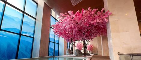 A large indoor space with a view of mountains through large windows, a central artificial cherry blossom tree, and a glass table with white pebbles.