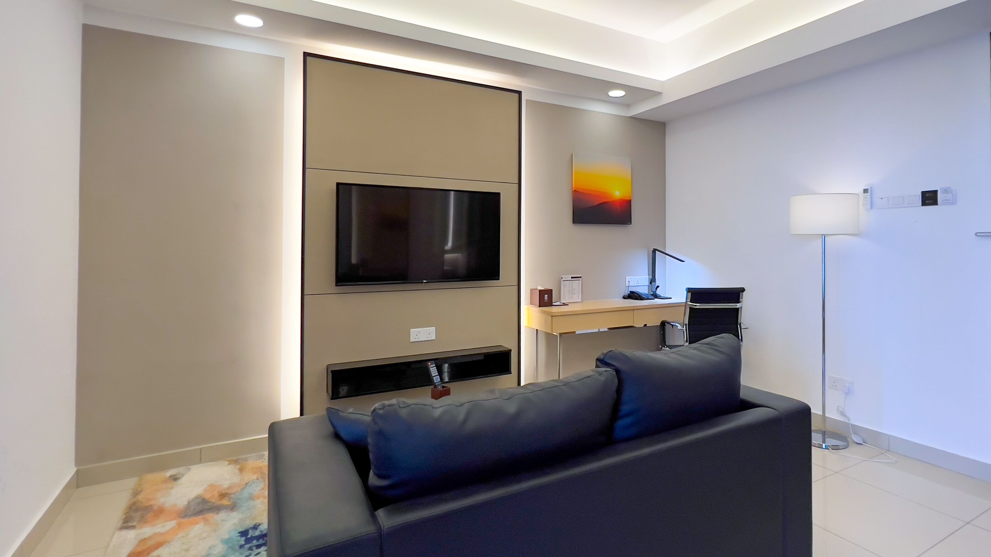 A modern living room with a flat-screen TV, a desk with a lamp, and a sofa.