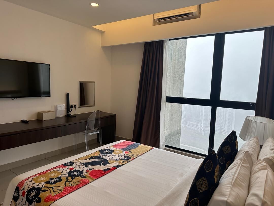 A hotel room with a bed, a desk with a flat-screen TV, a chair, a window with curtains, and a lamp.