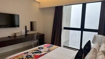 A hotel room with a bed, a desk with a flat-screen TV, a chair, a window with curtains, and a lamp.