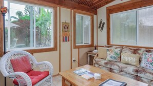 Una habitación acogedora con techo de madera, una silla de mimbre con cojín rojo, un sofá con almohadas tropicales y una mesa de madera con una revista y una foto enmarco.