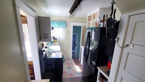 Une cuisine de taille réduite, équipée d’un réfrigérateur, d’une cuisinière et de placards. On peut apercevoir une table en bois et un tapis.