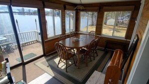 Un espace repas avec une table ronde et des chaises, de grandes fenêtres donnant sur un plan d’eau, et une terrasse en bois à l’extérieur.