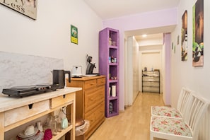 A compact kitchen with a marble countertop, wooden cabinets, a purple shelving unit, and a seating area with floral cushions.