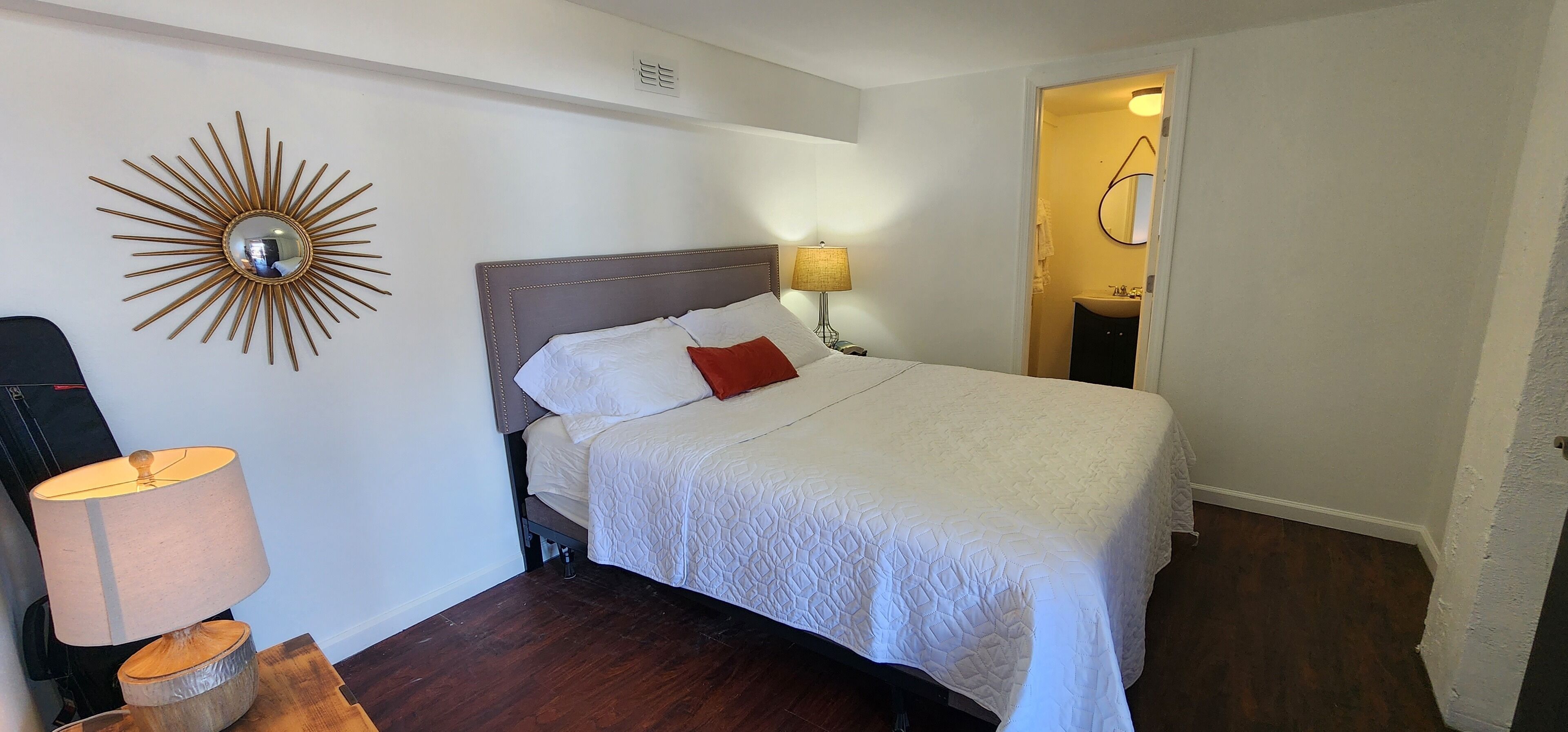 A neatly made bed with a white quilt, a single red pillow, and a wooden headboard. A round mirror with a sunburst design hangs on the wall above the bed. A lamp with a beige shade is on a wooden nightstand beside the bed.