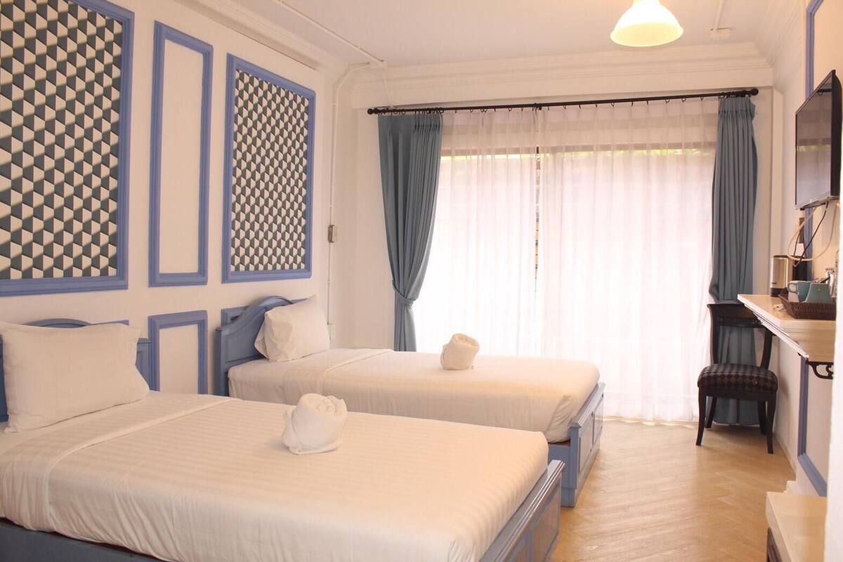 Een hotelkamer met twee bedden, een bureau, een stoel en een groot raam met gordijnen.