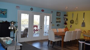 A dining area with a wooden table and white chairs, a blue wall with a turtle painting, and a door leading to an outdoor patio. - Dewey Beach Ocean Block Condo Elevator Parking Sleeps 10 (Dewey Beach)
