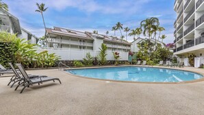 A swimming pool with lounge chairs, surrounded by palm trees and a building with balconies. - Ocean-View Condo in the Heart of Kona | Walk to Shops, Beaches & Dining (Kailua-Kona)