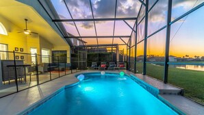 A swimming pool with a glass enclosure, a patio area with seating, and a view of the sunset over a body of water. - Single-Story Pool Home | Water Views & Game Room | Sleeps 11 (Kissimmee)