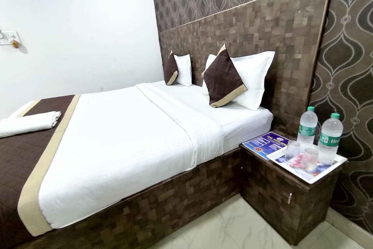 A neatly made bed with white linens, a brown headboard, and two pillows. There is a small wooden nightstand with a magazine and two water bottles.