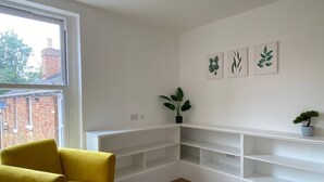 Une pièce avec une chaise jaune, des étagères blanches, une plante en pot et une œuvre d’art représentant des feuilles, encadrée et accrochée au mur.