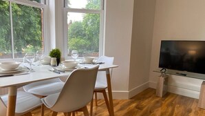 Un coin repas avec une table dressée pour quatre personnes, un téléviseur posé sur un meuble et une fenêtre donnant sur des arbres.