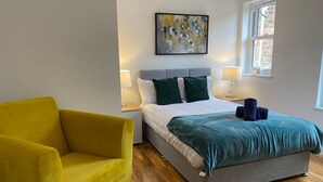 A bedroom with a bed, a yellow armchair, a painting on the wall, and a window.