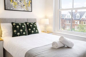 A neatly made bed with white bedding and two green pillows with white floral patterns, a rolled white towel, a bedside lamp, and a window with a view of houses.